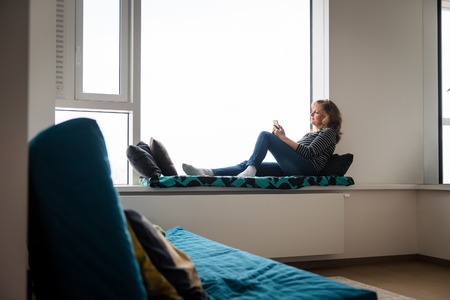 Mature woman sitting on window ledge bedの写真素材