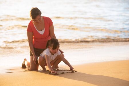 Woman guiding her daughter to draw beach heart on sandの写真素材
