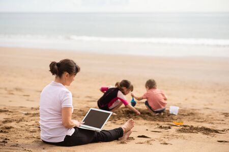 Busy mom teleworking on laptop while children playing beside shoreの写真素材