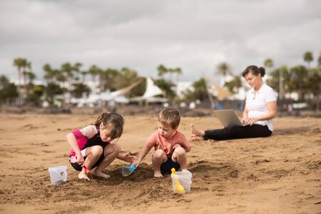 Children playing with beach toys while mother balances work-lifeの写真素材