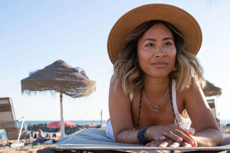 Beautiful asian girl on the beach playing with the sand and holding a summer sombrero hat at sunset facing the Atlantic ocean in Lisbon, Portugal. Object detail. Woman lifestyle concept of freedom.の写真素材