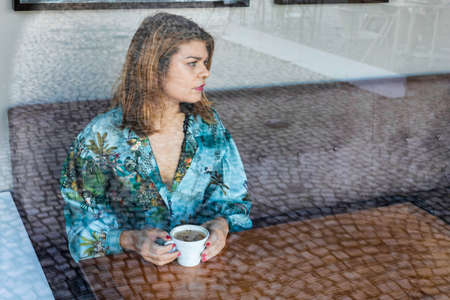 Portrait of a young woman sitting at the cafe and enjoying cup of coffee. The coffee bar is next to...