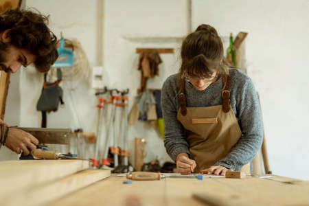 A young couple of carpenter working together in a small wood laboratory using a drill machine for timber. Couple crafting new home furniture in a carpentry workshop. young entrepreneurs concept A young couple of carpenter working together in a small wood laboratory using a drill machine for timber. Couple crafting new home furniture in a carpentry workshop. young entrepreneurs concept