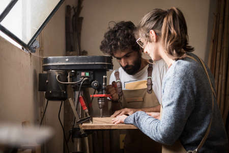 A young couple of carpenter working together in a small wood laboratory using a drill machine for timber. Couple crafting new home furniture in a carpentry workshop. young entrepreneurs concept A young couple of carpenter working together in a small wood laboratory using a drill machine for timber. Couple crafting new home furniture in a carpentry workshop. young entrepreneurs concept