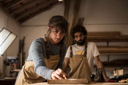 A young couple of carpenter working together in a small wood laboratory using a drill machine for timber. Couple crafting new home furniture in a carpentry workshop. young entrepreneurs concept A young couple of carpenter working together in a small wood laboratory using a drill machine for timber. Couple crafting new home furniture in a carpentry workshop. young entrepreneurs concept