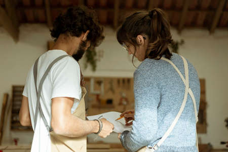 A young couple of carpenter working together in a small wood laboratory using a drill machine for...