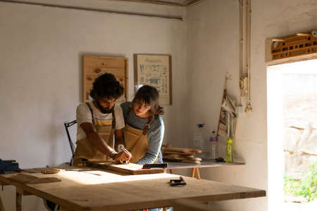 A young couple of carpenter working together in a small wood laboratory using a drill machine for...