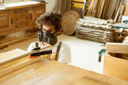 A young man working as carpenter in his wood workshop. Wood worker designing and handcrafting new house furniture using a piece of timber and wearing a full face mask for the dustの写真素材