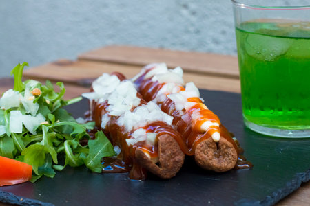 American junk food in a simple composition. crispy Fried sausages or hot dog with mayo and ketchup sauce and chopped onion. Some vegetables around like salad and tomatoes on a wooden background.の写真素材