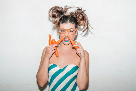 Beautiful cute lovely attractive funny happy smiling girl clothed in fashionable bikini are having fun playing with some carrots vegetables. Isolated on white background in summer hot colors editing.の写真素材