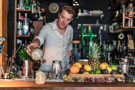 View of a professional bartender pouring a cocktail from a glass jar into a glass with lemon and ice, view of a small cocktail bar with fruits on counter, Lisbon, Portugal.の写真素材