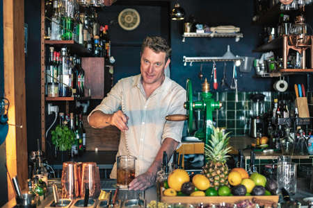 An expert bartender mixing a cocktail in a glass mixer with a long spoon, standing behind the counter of his beautiful cocktail bar in a night club.の写真素材