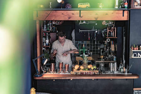 An expert bartender mixing a cocktail in a glass mixer with a long spoon, standing behind the counter of his beautiful cocktail bar in a night club.の写真素材