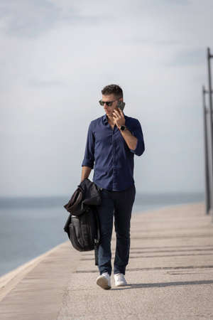 Beautiful and attractive young middle aged man walking alone and using the smart phone along the Lisbon riverside. Classy young entrepreneur wearing formal outfit. Successful lifestyle conceptの写真素材