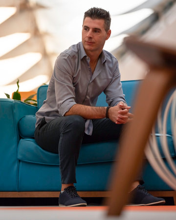 A young attractive freelance architect wearing a formal outfit and sitting on a blue couch inside a shopping mall in Lisbon, Portugal. Smiling young man with elegant dress working remotely worldwide.の写真素材