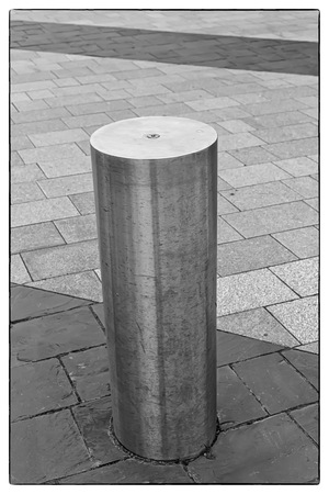 Black and white photo of a Stainless Steel bollard on grey pavement with a few lines of dark grey...