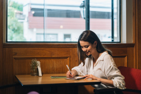 Student writing what she has learned in the library. Portrait of a beautiful and intelligent young woman smiling while writing in a notebook on the table at homeの写真素材