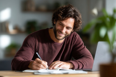 Smiling man writing in notebook while working on laptop. Concept of creativity, productivity, and work-life balance.の素材