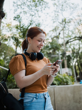 Young woman checks messages on her smartphone outside with headphones. Modern lifestyle and digital...