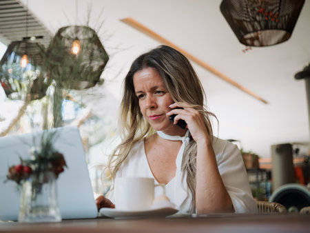 Professional woman talking on the phone while working on a laptop in a cafe. Business communication and multitasking concept.の写真素材
