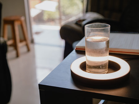 Photograph of a glass glass with water on a black table, illuminated by warm light, with an...