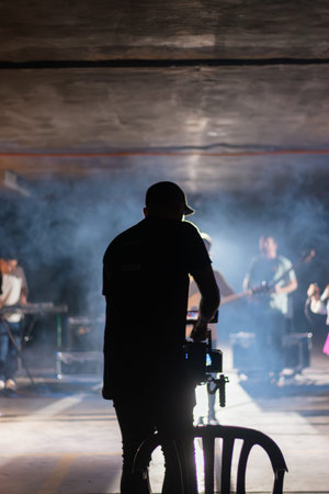 A man stands in front of a group of musicians, filming them. The scene is dimly lit, with smoke in the background. Scene is mysterious and somewhat ominousの写真素材