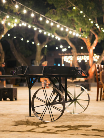 A black cart with a wheel is sitting in a courtyard. The courtyard is lit up with lights, creating a...