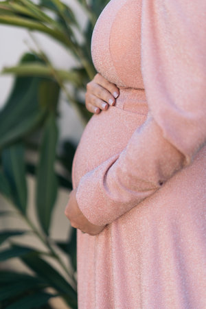 A pregnant woman is wearing a pink dress and is leaning on a plant. Concept of comfort and...