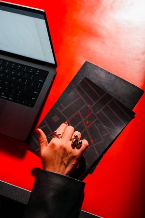 Hand rests on dark map atop vibrant red surface. Laptop sits beside map, suggesting planning or navigation. Fingered hand wears rings, adding personal touch to the sceneの写真素材
