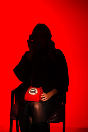 Red glow illuminates silhouette of a person. They hold phone close to their ear. Red bag rests on their lap. Seated, they appear engaged in conversation. Dramatic lighting adds mystery to the sceneの写真素材