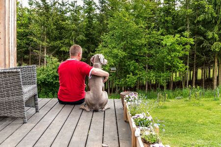 man is sitting with a dog on the terrace of the house and looking into the woods.の写真素材
