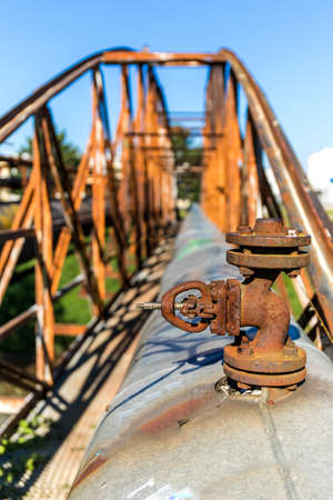 Old rusty iron bridge over the river with a steam pipeline. Heat supply. Industrial equipment. Detailed view of a steam pipeline valve.の写真素材