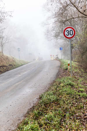 Fog on a country road. Speed traffic sign. Dangerous place. Slippery road. Foggy day. Empty road in...