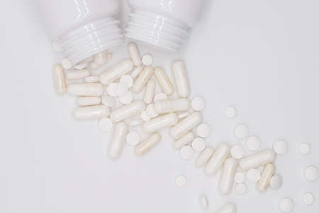Heap of white pills and capsules, medicines, vitamins and useful food supplements scattered from sterile container on a white light background, flat lay with blank space top viewの写真素材