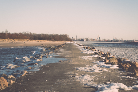 frozen beach near shipyard and sea port with rays of sun and wavebreaker - vintage retro grainy film effectの写真素材