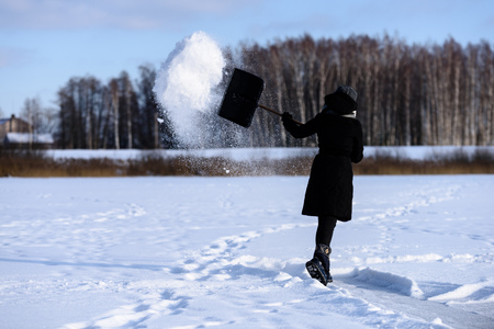 young woman in black coat cleaning ice from snow on a frozen lake to make skating ground. throwing snow in the airの写真素材