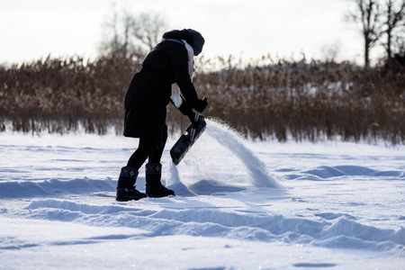 young woman in black coat cleaning ice from snow on a frozen lake to make skating ground. throwing snow in the airの写真素材
