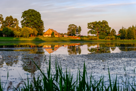 countryside scene by the lake at sunset with trees over the pond and house on the shore. dramatic colored skyの写真素材
