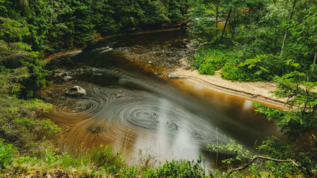 long exposure rocky mountain river in summer with high water stream level in forest with trees and sandy foreground shoreの写真素材