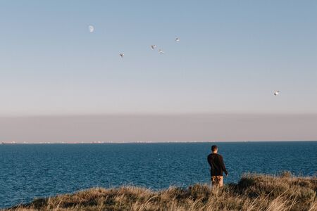 Sunset in the Crimea. A man stands on the shore and admires the sea. Seagulls and the moon are visible in the sky. Cargo barges at sea. Copy spaceの写真素材