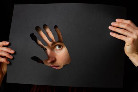 A young adult  woman looking through hand shaped hole in black paperの写真素材