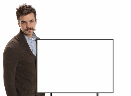 Attractive young man with a handlebar mustache and a brown sweater stands in front of a blank white sign with black border, isolated on whiteの写真素材