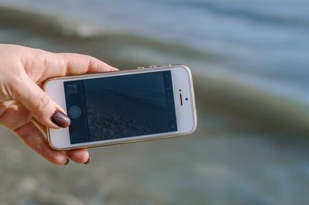 Woman hand with manicure holding white mobile phone with wave and sea in background.の写真素材