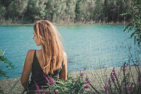 A beautiful girl stands with her back to the camera and face the river. Turquoise water, plants. Summer. Landscape.の写真素材