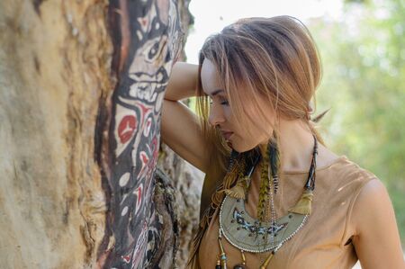 A beautiful dark-haired girl in the boho style stands by a tree. Beautiful nature, summer. Ethnic jewelry.の写真素材