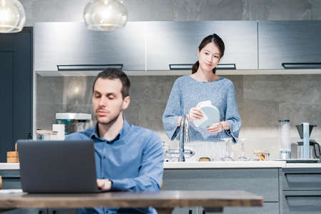 A man working at a dining table and a woman washing dishes in the kitchenの写真素材