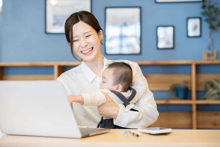 Asian woman holding a baby and operating a laptop indoorsの写真素材