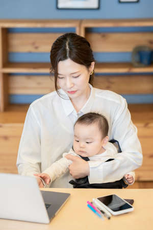Asian woman holding a baby and operating a laptop indoorsの写真素材