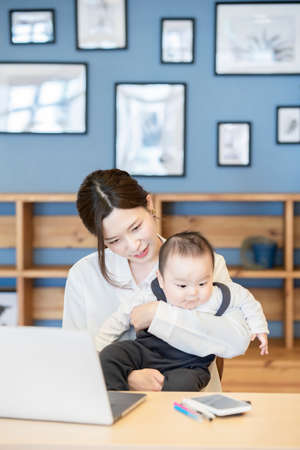 Asian woman holding a baby and operating a laptop indoorsの写真素材