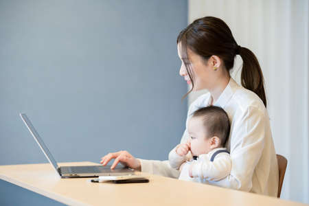 Asian woman holding a baby and operating a laptop indoors
